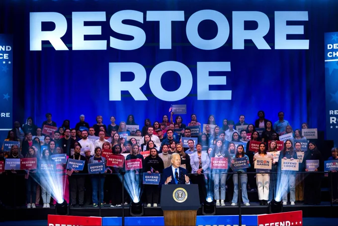 A photo of President Biden delivering remarks at a reproductive rights event. Behind him are supporters holding signs that read "Biden-Harris" and "Defend choice" as well as a large sign that reads "Restore Roe"