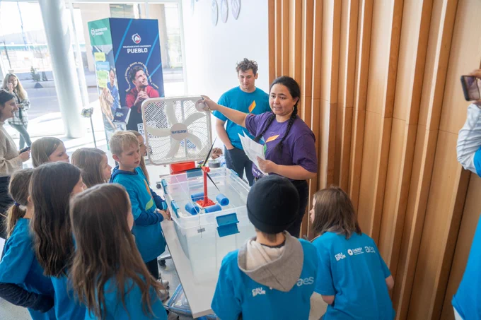 Group of kids surrounding a fan on a table with a bucket of water in front