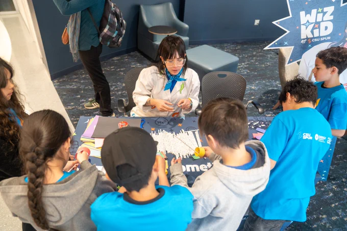 Young kids surround a table making straw planes with Wiz Kid