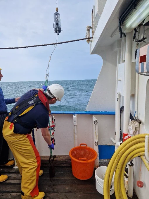 The image shows a fisherman in yellow protective gear holding a fishing line on the deck of a boat. He is holding the line at the bottom and beside him is an orange storage basket.