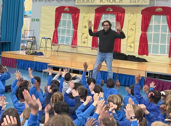 Our Patron of Reading Sav Akyüz stands in front of a class of primary school students with his arms raised as a character.