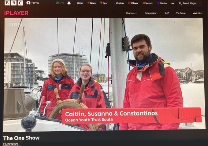 Screengrab from BBC One Show of our team members in red oilskins on board Prolific, by the wheel with mizzen mast behind them, with BBC branding and a caption reading"Caitlin, Susanna & Constantinos, Ocean Youth Trust South".