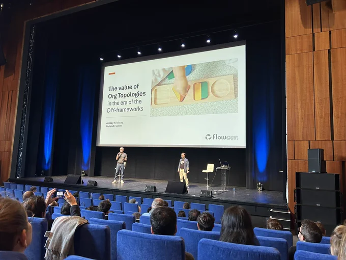 Two speakers on the stage of flow con. Behind them their first slide « the value of org topologies in the era of the DIY frameworks »