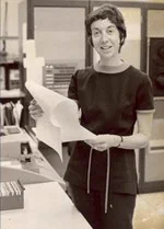 Judith (Levenson) Clapp stands holding a printout in front of computer equipment.