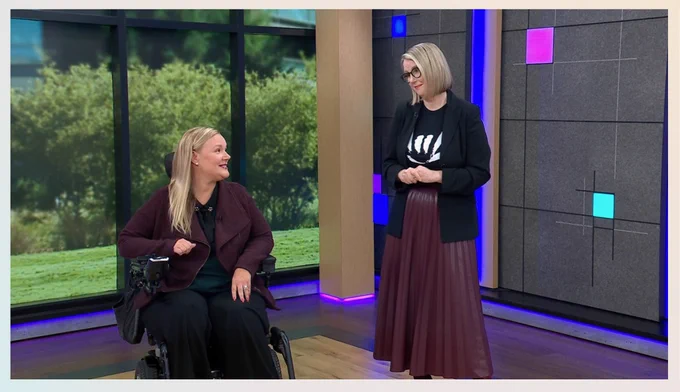 Jessica in power wheelchair looks at Jenny on a stage with trees outside the window