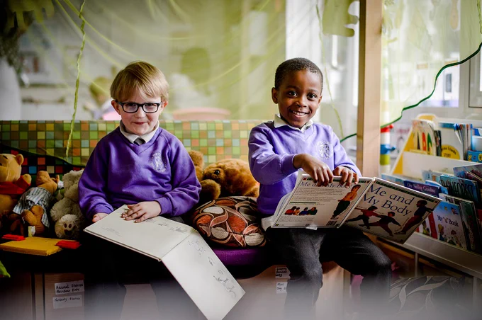 Two primary schools children sitting in a colourful library reading a book and smiling to the camera.