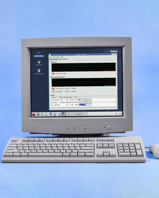 A computer that might have been manufactured in the early 2000s is shown. There is a large monitor with a white-gray color, connected to a wide keyboard and mouse with a similar aesthetic. The monitor screen shows the desktop view of Fedora Core 1, the first release of Fedora Linux in November 2003. A process activity monitor application is open on the desktop.