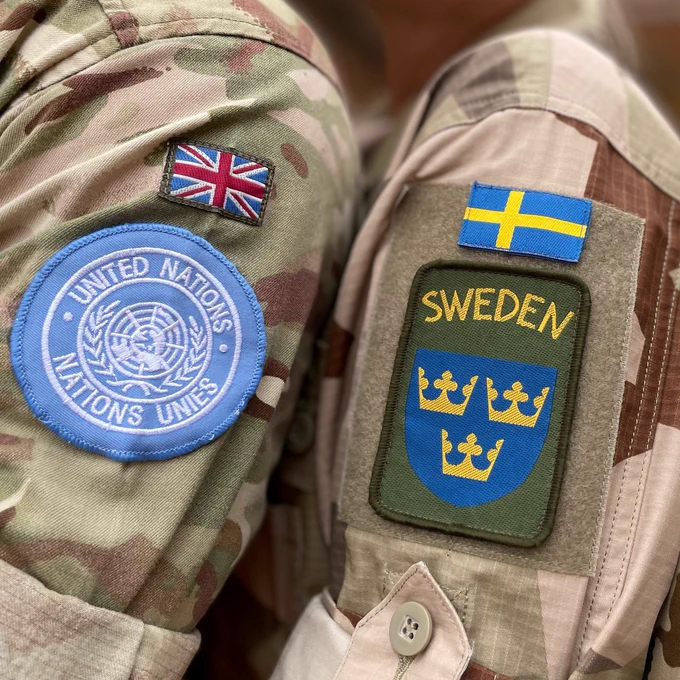 Two soldiers stand side by side with their respective flashes showing on their arms. The arm on the left shows the UK flag with a United Nations badge below. The arm on the right shows a Swedish flag with a Swedish badge below.