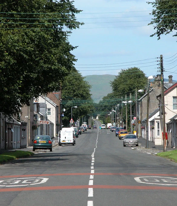 Today's 'Where in Scotland' image. It is a vertical image showing a view along a village street with buildings on both sides and lots of trees. There are multiple lines of wires crossing overhead the road. The scene is in sunlight.
