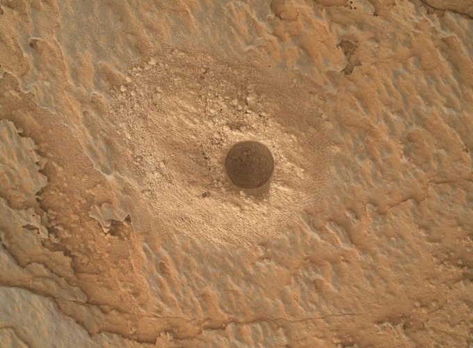 A color image shows a close-up shot of the Curiosity Mars rover's 40th successful drill hole named "Mineral King." The hole is in the center of the image and the detail shows the grains of the drilling that has piled up around the circle. The Martian surface is a light beige color and the rock surface surrounding the hole shows many layers and has almost a soft shine to it.