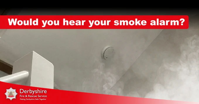 Image of a landing. The camera is facing the ceiling, close to the stairs, and there is a smoke alarm. Smoke is drifting up towards the alarm. Text in a red box at the top of the image reads 'Would you hear your smoke alarms?' At the bottom of the image is a red swoosh which features the Derbyshire Fire & Rescue Service badge