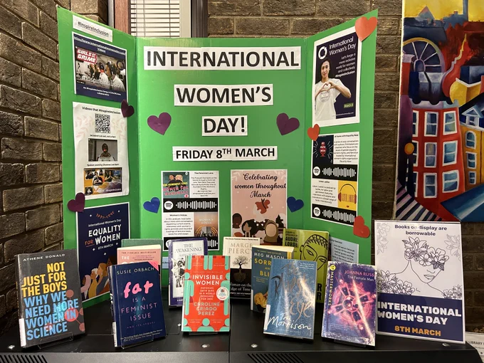 A green display board with International Women's Day! Friday 8th March in large print, surrounded by posters and information relating to the event. In front of the board there are a selection of books written by, or about women.