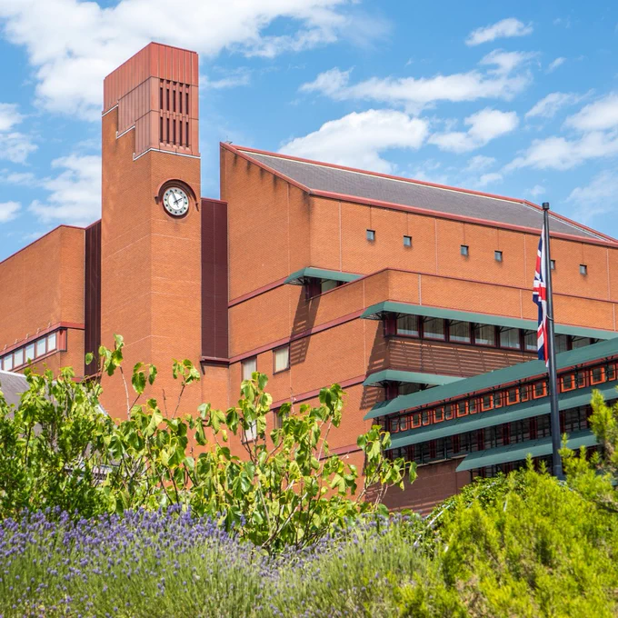 An exterior view of the British Library at St Pancras, showing the clock tower. Green plants and bushes frame the bottom of the photograph, and a blue sky streaked with white fluffy clouds is above.