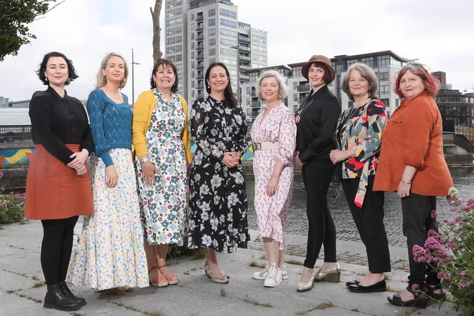 The recipients of the Markievicz Award in 2023 stand with Minister Catherine Martin. People in brightly coloured clothes stand in a row in an outdoor setting. They smile and look directly at the camera. Behind them a river and buildings are visible.