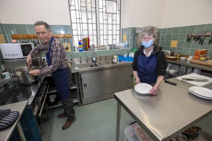 A community kitchen preparing for service. There is a woman holding plates and a man stirring a pot.