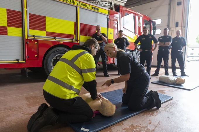 A fire station. There is a woman instructing a man on how to do CPR on a dummy. There is a fire truck and a group of firefighters stood behind them. 
