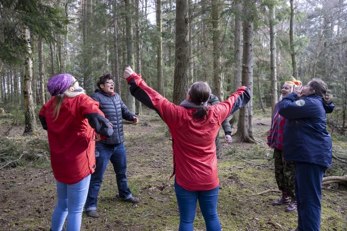 A group of women in a forest, they are obviously shouting and some are holding their arms in the air. They are releasing energy together.