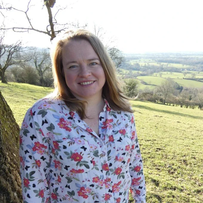 Head of Ecology Dr Caroline Bulman stood next to a tree, smiling at the camera, with fields and countryside in the background
