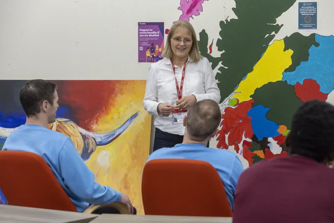 A woman wearing a lanyard is presenting to group of seated men. There is a colourful mural of the Scottish map on the wall behind.