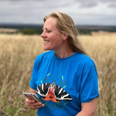 Senior Surveys Officer Dr Zoë Randle holding a phone while sat on a bench, wearing a blue t-shirt featuring a Jersey Tiger moth, with a meadow behind
