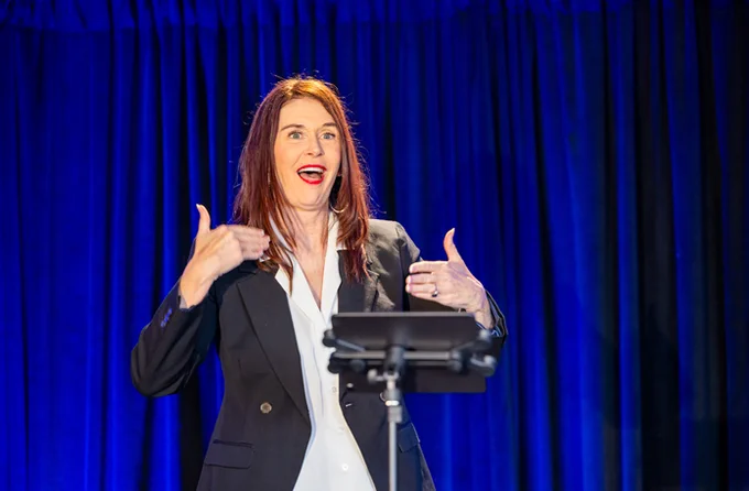 AnnMarie Killian, a white woman with long brown hair, is signing in American Sign Language (ASL). She wears a white blouse and a black suit, complemented by a touch of red lipstick. The photo takes place at the TDI Diamond Gala.