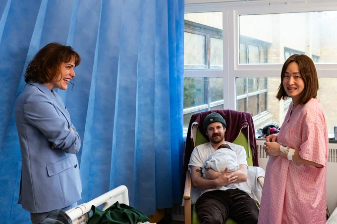 Two women, one in a hospital gown and one in a suit, and a man sitting down with a newborn baby in his arms. 