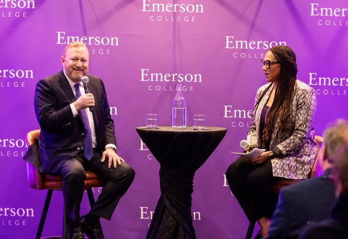 A person with a microphone sits and converses with another person at a tabletop.