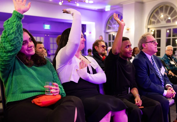 Seated people raise their hands and listen to speakers.