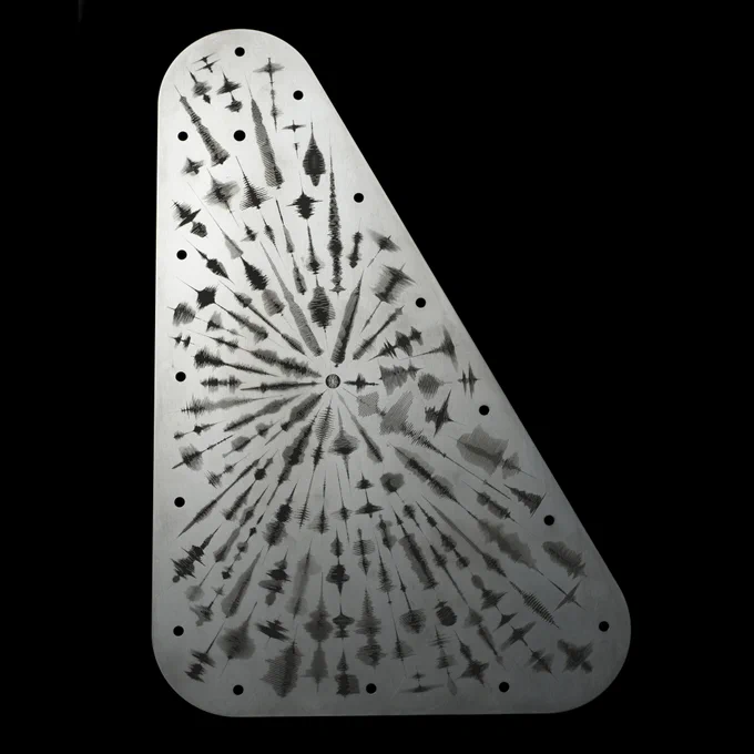 A full view of the Europa Clipper vault plate displaying an intricate pattern of waveforms for the word 'water' in over 100 languages, each emanating from the center to the edges. The waveforms present as audio frequency waves. The vault plate is metal silver and the engravings are black, set against a black background. 