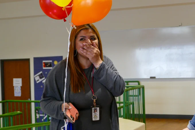 An educator places a hand over her mouth in surprise as she holds a set of three balloons. 