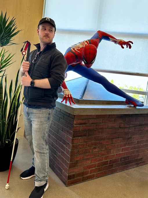 James stands to the left, holding a white cane with his right hand, indicating his visual impairment. Positioned behind him is a life-size statue of Spider-Man, posed dynamically on a brick platform. The setting appears to be an indoor space with large windows allowing natural light, and there are plants in the vicinity.