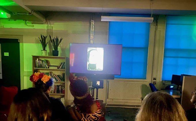A room with blue windows and a green light on left, all focus is on the Tv screen in middle as the premiere of Sheila’s film is shown.  image on tv is an old leaflet for positively women drawn and copied roughly as it was back then, audiences head in foreground