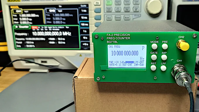 A small green FA-2 frequency counter sitting on top of a cardboard box. The screen reads 10000000.000 Hz. In the background, a Rigol function generator in frequency counter mode, measuring the same signal and showing 10.000,000,000,0 Mhz