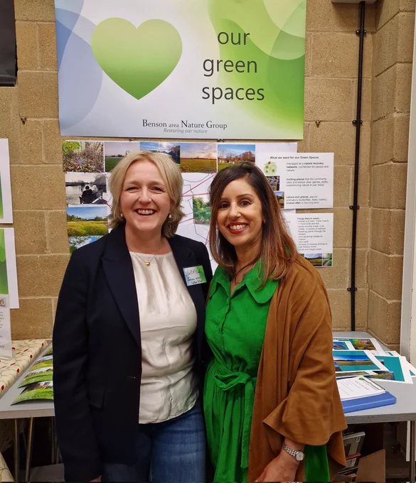 2 women (Edel and Geeta) at the Benson Nature event standing in front of a nature display on Benson. Poster reads "Our Green Spaces". Poster includes various photos across the seasons of Benson nature. 