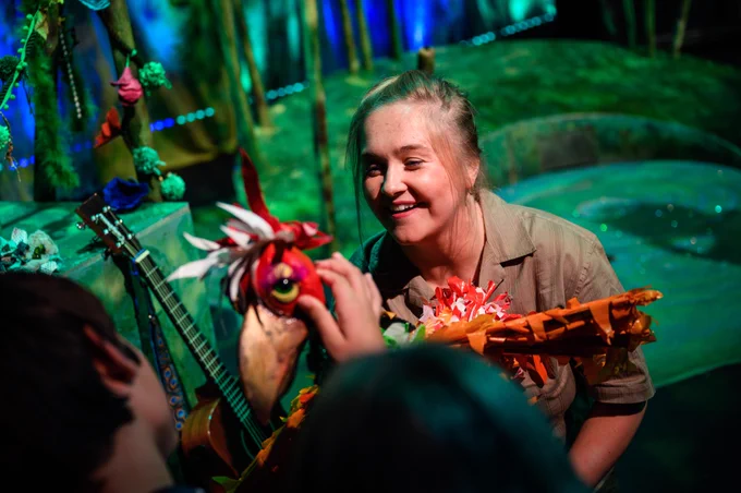 A person holds a puppet of a colourful bird and assists a child to explore it.