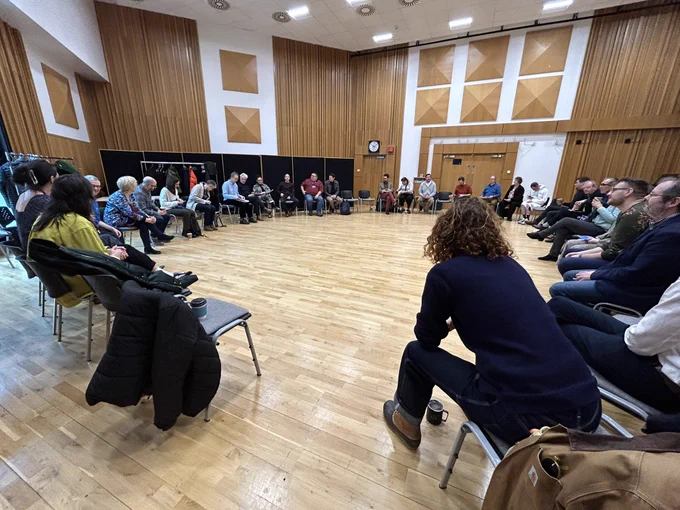 A large room with a pale wood floor and wood-clad walls. 45 people sit in a circle on grey and black chairs.