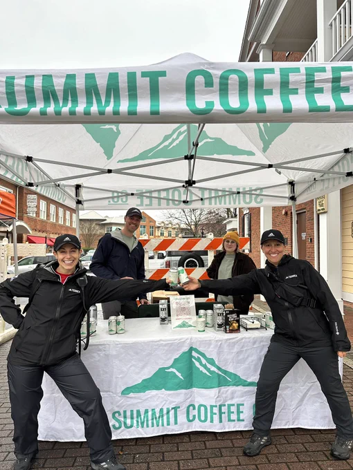 Two Atlanta Track Club staff members cheers a can of Summit Coffee, and two Summit Coffee employees smile at their table and tent at the Members 10K.