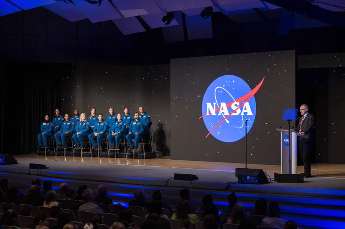 On the right side of a stage, NASA Associate Administrator Jim Free gives a commencement speech addressing the 10 new NASA astronauts and 2 UAE astronauts sitting on the left side of the stage during the 2024 NASA Astronaut Graduation ceremony held in the Teague Auditorium at NASA's Johnson Space Center.