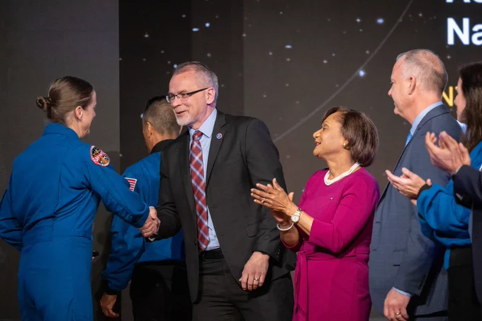 NASA Associate Administrator Jim Free smiles and shakes hands with new NASA astronaut Jessica Wittner. Johnson Space Center Director Vanessa Wyche claps while Flight Operations Director Norm Knight looks on.