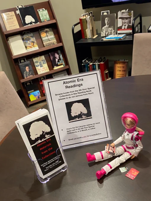 Photo of table with sign and brochure for Atomic Era display; astronaut Barbie sitting nearby reading several books on history of the Atomic Age. Books relating to history of this period on shelves in the background.