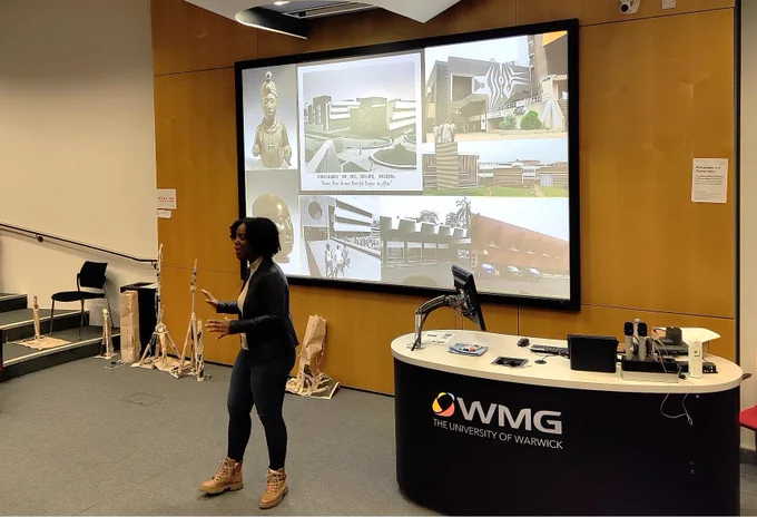 Yewande giving her presentation standing at the front of an auditorium, behind her on the screen are images of Nigerian culture and buildings.