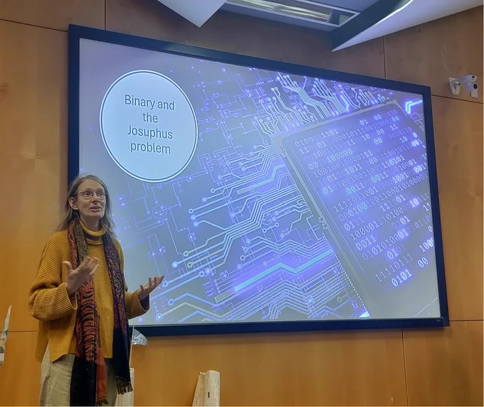 Helena standing in front of a screen which shows a printed circuit board and binary images with the text Binary and the Josuphus problem. 
