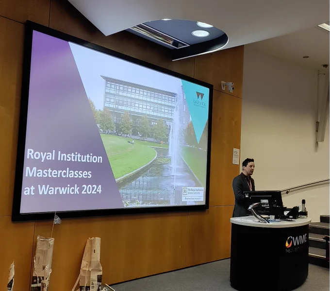 Phil giving a presentation standing behind a lecture podium.  Behind him is a screen showing the text Royal Institution Masterclasses at Warwick 2024.