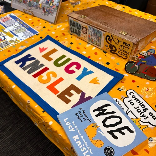 A table with a bright yellow table cloth and a small banner reading "Lucy Knisley" in colorful letters. We can also see an early copy of her next book "Woe," featuring comics about her orange cat Linney, with a small sign saying "Coming out in July!" Also visible are an art box and several prints.