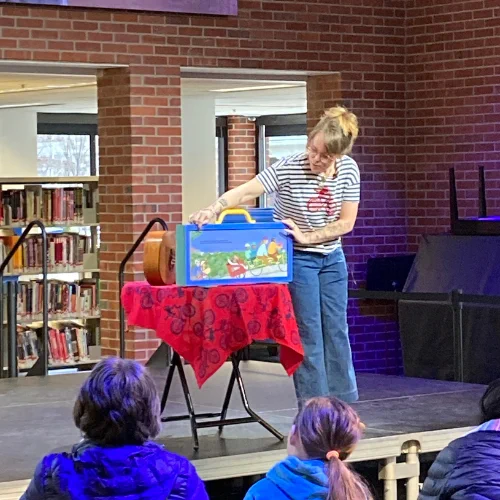Lucy standing beside a bright blue box with a yellow handle, in the middle of which is a page from Ride Beside Me mounted on a board. She is preparing to turn the page by changing out the board.