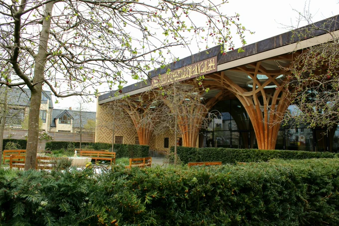 The Islamic Garden at the front of Cambridge Central Mosque on an overcast day. An octagonal stone fountain is surrounded by curved oak benches, where visitors can sit and enjoy the sound and movement of the water, the colours and fragrances of the flowers, and the shade of the trees.