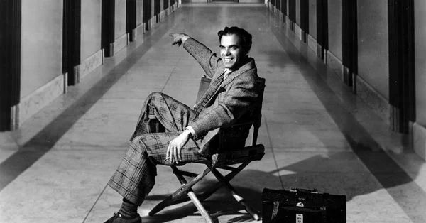 A black and white still from the film Frank Capra: Mr America. A white man in a plaid suit sits in a director's chair in a long, marble-floored hallway, grinning and holding out his arms.