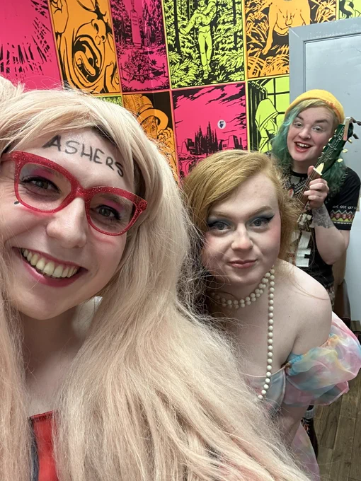 Céilí, Rose, and Ash Jones standing and smiling together in a green room. Ash is holding a guitar, Rose has a pearl necklace on, and Céilí has ASHERS written on her forehead