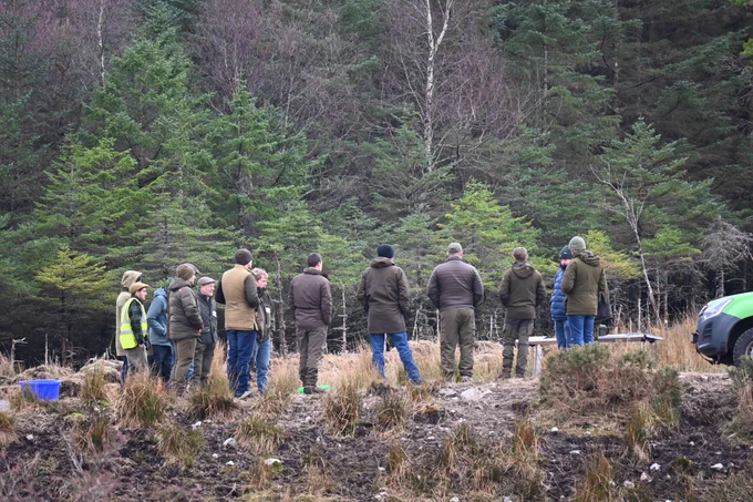 Group of people standing in a circle outside, listening to instructor, green trees in background