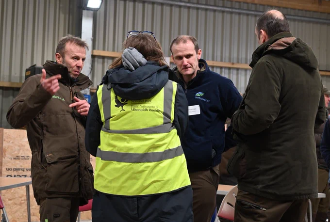 Two NatureScot staff members talking  to two people. Image is taken indoors in large shed-type building with corrugated walls.